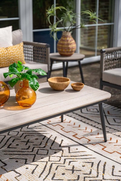 Heller, organisierter Loungebereich im Freien mit Couchtisch in Holzoptik, Gartenstühlen und einem gemusterten Outdoor-Teppich.