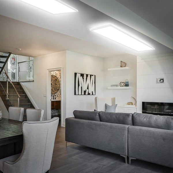 Modernes Wohnzimmer mit zwei flachen LED-Deckenpanelen, großem grauen Sofa, Esstisch und offener Treppe in heller Atmosphäre.