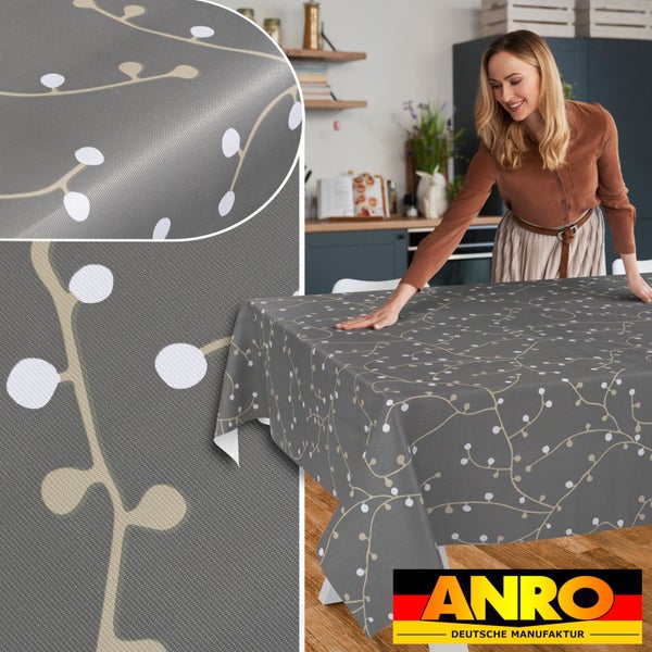 Eine Frau breitet eine gemusterte Tischdecke mit Zweigmotiv auf einem Küchentisch aus. Detailansicht von Gewebe und Muster. ANRO Logo.