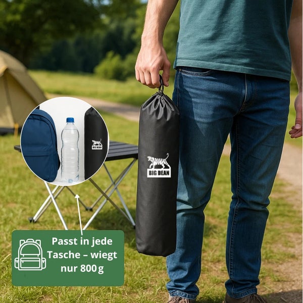 Person hält eine schwarze Tragetasche mit dem Big Dean Logo in der Hand, Campingausrüstung im Hintergrund.