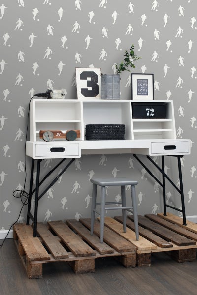 Szene mit einem Schreibtisch, einem Hocker und dekorativen Gegenständen vor einer Tapete mit Fußballmotiven.