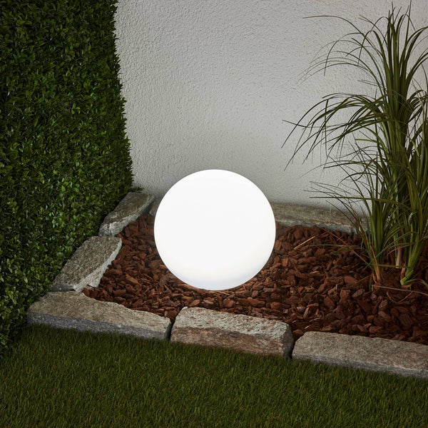 Leuchtende LED-Kugelleuchte in einem Gartenbeet mit Rindenmulch, eingefasst von Natursteinen, neben Gräsern und einer Hecke.
