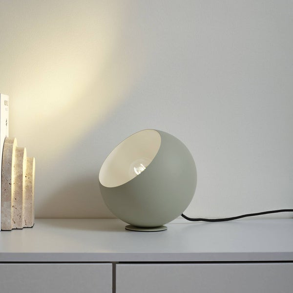 Moderne kugelförmige Tischleuchte mit schräger Öffnung auf einem Sideboard neben dekorativen Steinobjekten in hellem, minimalistischem Ambiente.