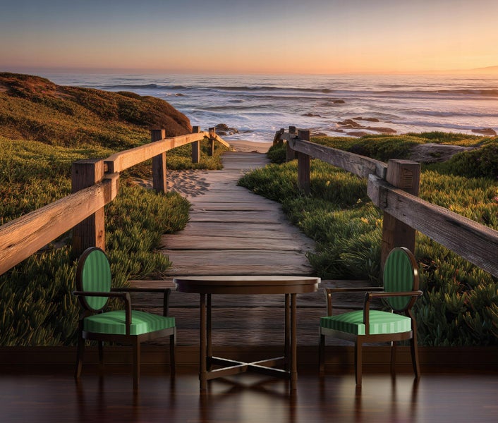 Fototapete mit Küstenlandschaft bei Sonnenuntergang, Holzsteg zum Meer, davor ein runder Holztisch und zwei grün gestreifte Polsterstühle.