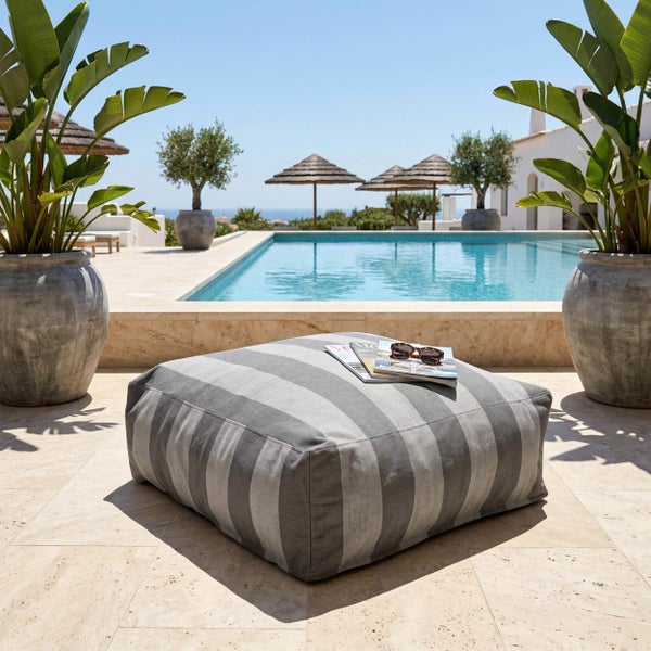Gestreifter quadratischer Outdoor-Sitzsack auf einer hellen Steinterrasse an einem Swimmingpool mit großen Pflanzen in Kübeln.