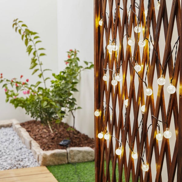 Solar-Lichterkette mit warmweißen Kugelleuchten an einem dunklen Holzspalier in einem Garten.