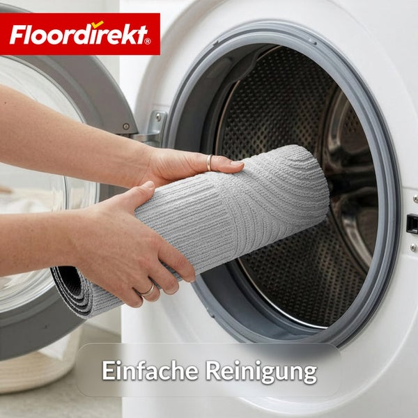 Zwei Hände legen eine zusammengerollte graue Badematte in eine weiße Waschmaschine zur Reinigung. Floordirekt Logo.