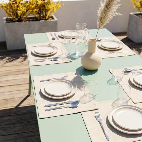 Gedeckter Gartentisch in Mintgrün auf einer Holzterrasse mit beigen Platzsets, weißen Tellern, Besteck und einer Vase mit Pampasgras.