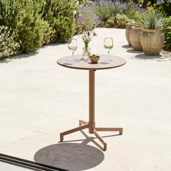 Runder Bistro-Gartentisch auf einer Terrasse mit Weingläsern, Blumen und Oliven, umgeben von Pflanzen und großen Töpfen.