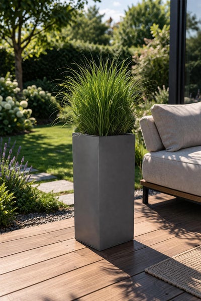 Hoher quadratischer Pflanzkübel mit Ziergras auf einer Holzterrasse neben einem Gartensofa im hellen Außenbereich.