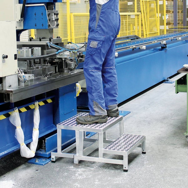 Aluminium-Arbeitstritt mit zwei Stufen und rutschhemmender Oberfläche in einer industriellen Werkshalle neben einer Maschine.