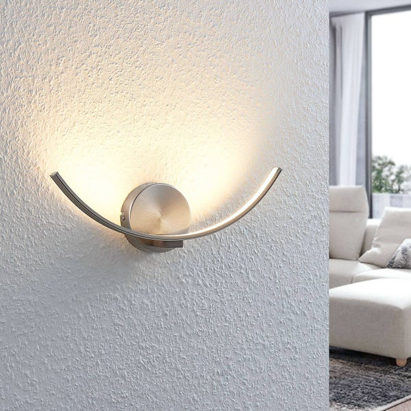 Moderne LED-Wandleuchte aus Metall in geschwungener Form an einer strukturierten Wand in einem hellen Wohnzimmer.