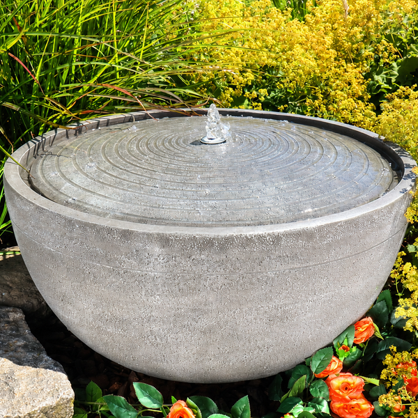Halbkugelförmiger Gartenbrunnen in Steinoptik mit konzentrischem Rillenmuster auf der Oberseite und mittigem Wasseraustritt in einem Garten.