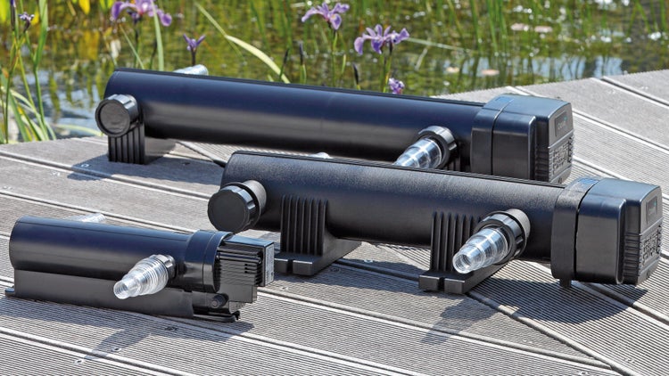 Drei Oase UVC-Vorklärgeräte in verschiedenen Größen auf einem Holzsteg an einem Gartenteich mit lila Blumen.