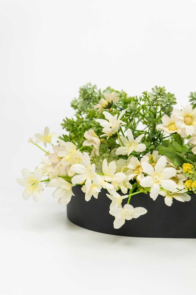 Künstliches Blumengesteck mit weißen Blüten und grünem Blattwerk in einer schwarzen Schale.