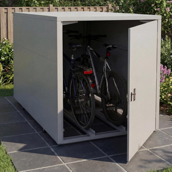 Fahrradbox mit geöffneter Tür und zwei Fahrrädern im Inneren, auf einer Steinterrasse im Garten neben einem Holzzaun.