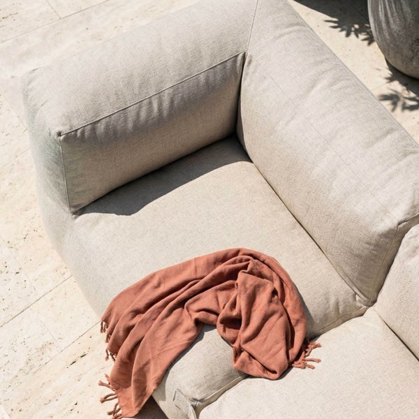 Beiges Outdoor-Lounge-Sofa aus strukturiertem Stoff mit einer terrakottafarbenen Fransendecke auf einer Steinterrasse.