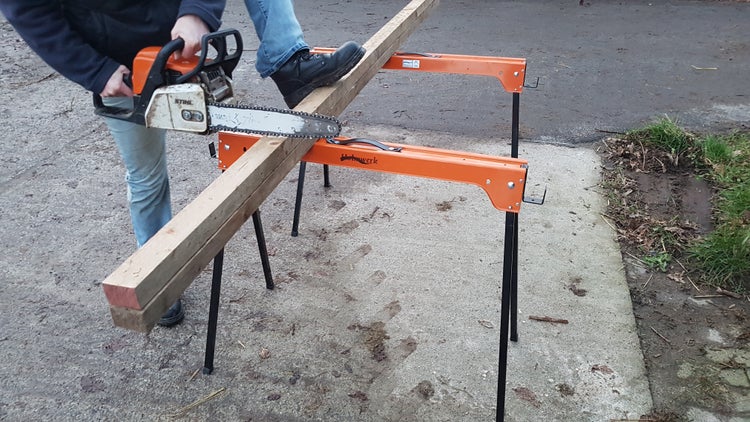 Eine Person sägt ein Holzbrett mit einer Kettensäge auf einem Sägebock.