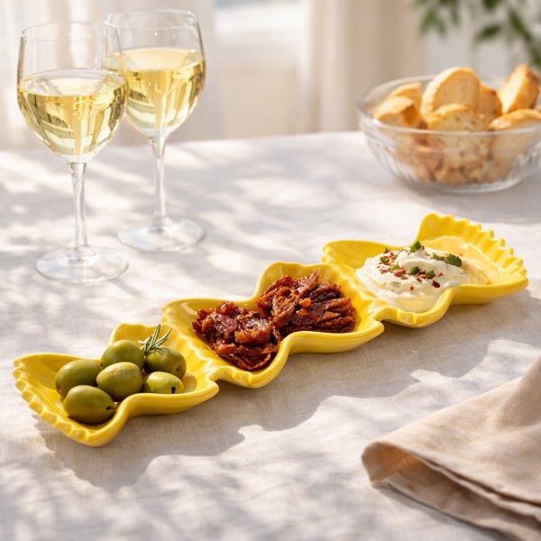 Gelbe Servierschale aus Keramik in Wellenform mit Oliven, getrockneten Tomaten und Dip auf einem gedeckten Tisch mit Wein und Brot.