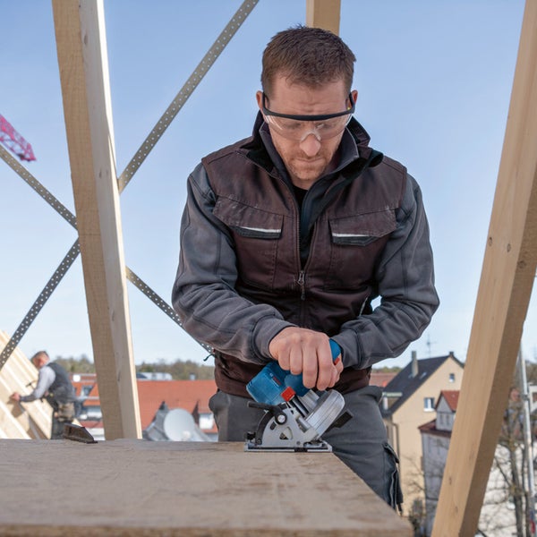 Handwerker mit Schutzbrille schneidet eine Holzplatte mit einer batteriebetriebenen Handkreissäge auf einer hellen Baustelle.
