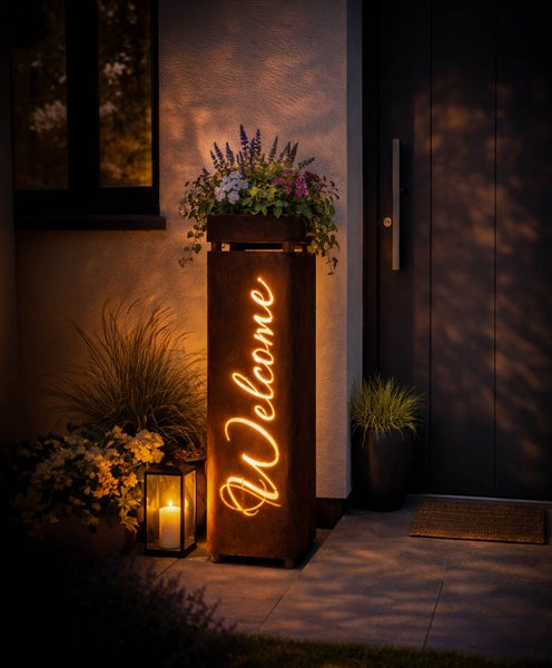 Beleuchtete Dekosäule aus Metall mit Welcome Schriftzug und Pflanzschale am Hauseingang in der Abenddämmerung.