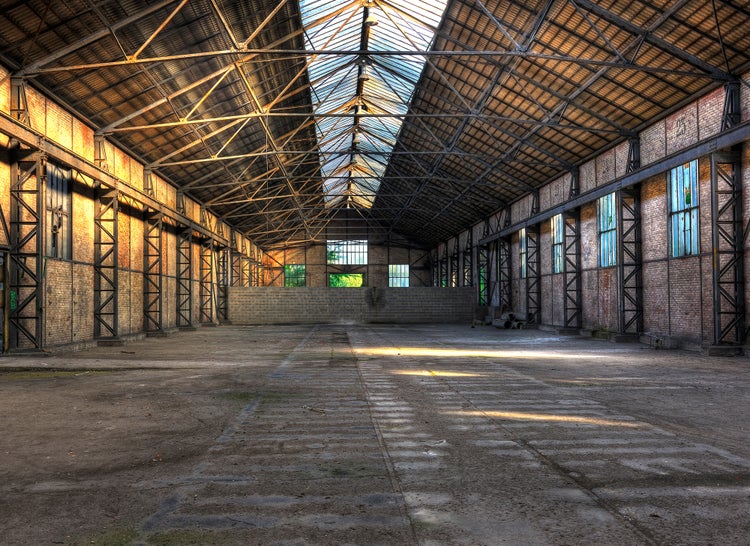 Große, leere Industriehalle mit offenliegenden Stahlträgern, Ziegelwänden und einem Glasoberlicht. Betonboden und natürlicher Lichteinfall.