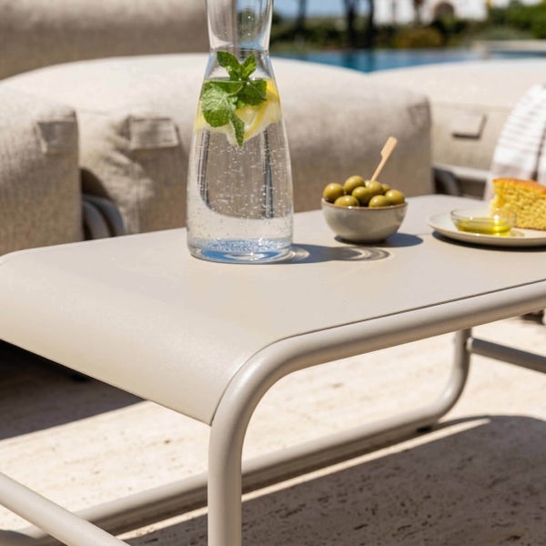 Moderner beigefarbener Outdoor-Loungetisch mit abgerundeten Kanten, dekoriert mit einer Wasserkaraffe und Snacks im hellen Außenbereich.