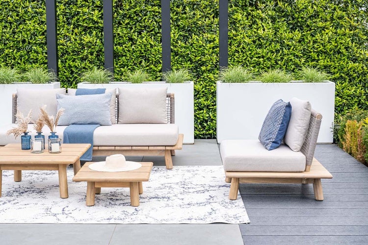 Modern gestalteter Außenbereich mit Lounge-Set aus Holz, Polstern, Couchtisch und Beistelltisch auf einer Terrasse vor einer Hecke.