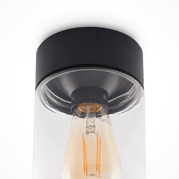 Nahaufnahme einer Leuchte mit schwarzem Sockel und einem transparenten zylindrischen Glasschirm mit einem LED-Filament-Leuchtmittel.