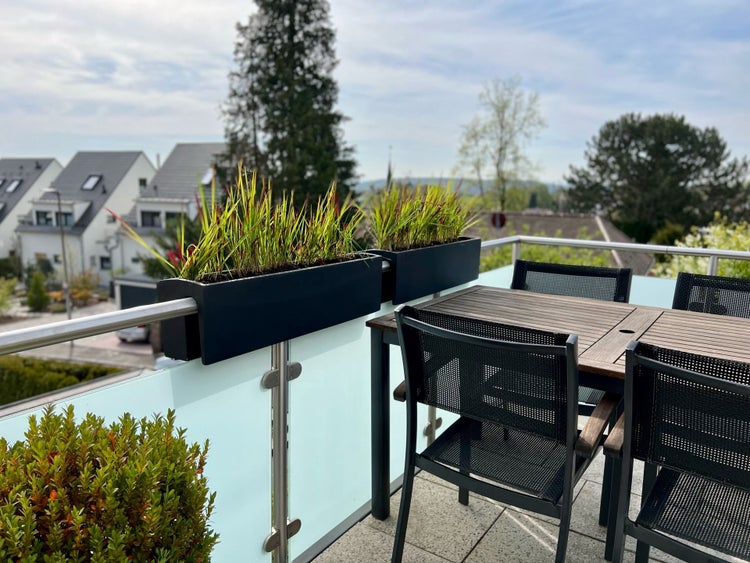 Balkongestaltung mit anthrazitfarbenen Blumenkästen am Geländer, bepflanzt mit Gräsern, sowie einem Gartentisch aus Holz und schwarzen Stühlen.