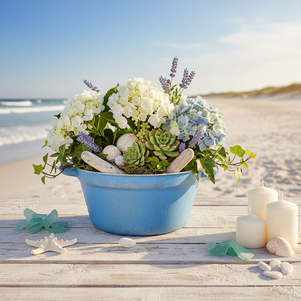 Maritimes Blumenarrangement mit Hortensien, Sukkulenten und Muscheln in einer blauen Schale auf einem Holztisch am Strand mit Kerzen.