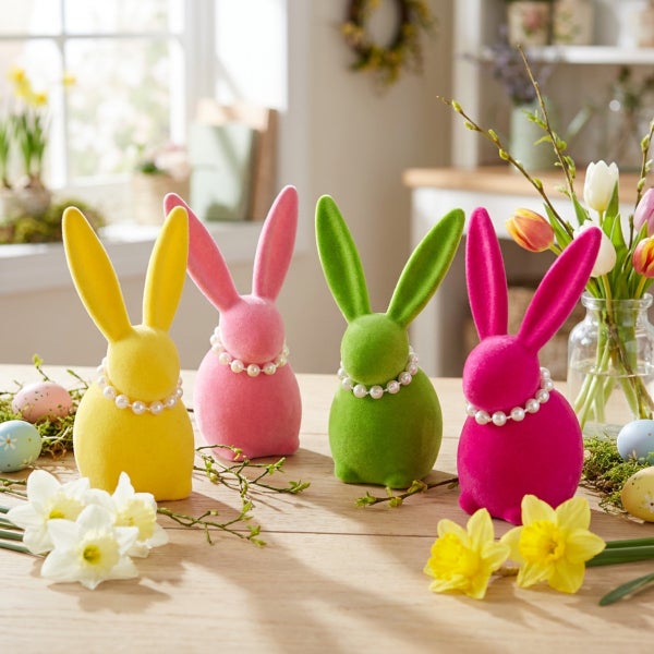 Vier flauschige Osterhasen Figuren mit Perlenketten als Frühlingsdekoration auf einem Holztisch mit Narzissen und Ostereiern.