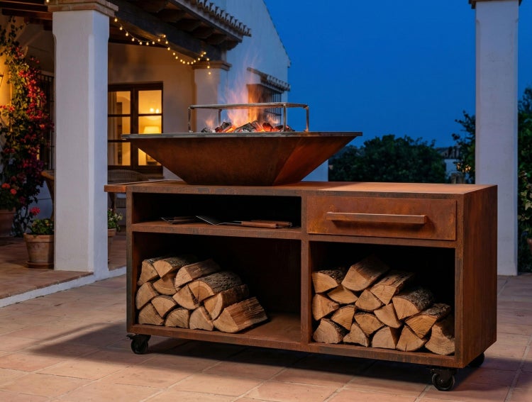 Mobile Außenküche aus Metall mit Feuerschale, Grillrost, Schublade und Holzlager auf einer Terrasse am Abend.