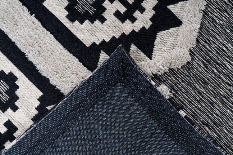 Nahaufnahme eines Teppichs mit schwarz-weißem geometrischem Muster und umgeklappter Ecke, die die strukturierte Rückseite zeigt.