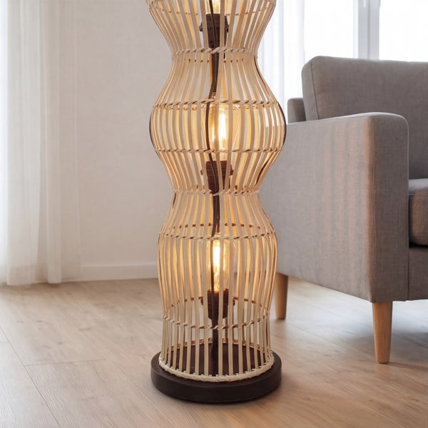 Stehleuchte aus geflochtenem Bambus in Sanduhrform mit sichtbaren Leuchtmitteln in einem hellen Wohnzimmer neben einem grauen Sofa.