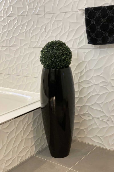 Hohe, glänzend schwarze Bodenvase mit grünem Buchsbaum-Kunstball in einem modernen Badezimmer mit weißem 3D-Wandbelag.