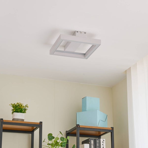 Quadratische LED-Deckenleuchte montiert in einem hellen Zimmer über Regalen mit Pflanzen und Dekorationsboxen.