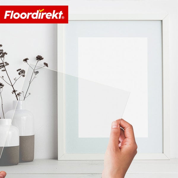 Eine Hand hält eine transparente Acrylglasplatte vor einen weißen Bilderrahmen. Floordirekt Logo.