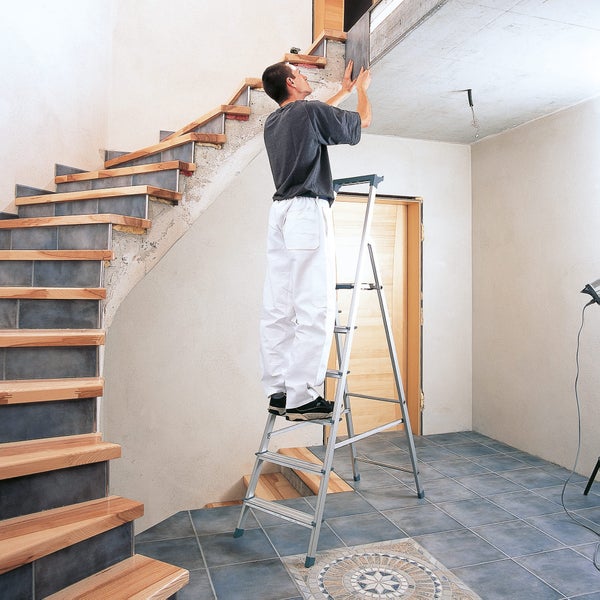 Mann auf einer Leiter, der eine Platte an der Decke befestigt. Im Hintergrund sind eine Treppe und Fliesen zu sehen.