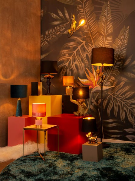 Dekorative Lampen in verschiedenen Formen und Größen auf farbigen Podesten in einem Zimmer mit Dschungeltapete und einem grünen Teppich