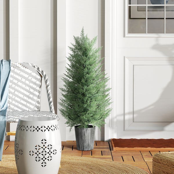 Künstliche Zypresse im grauen Topf auf einer hellen Terrasse mit Holzboden neben einem weißen dekorativen Hocker und einer weißen Haustür.