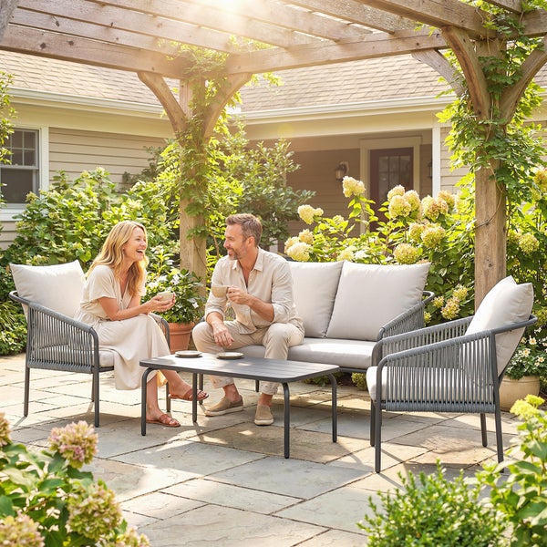 Moderne Gartenlounge unter einer Holzpergola mit Sofa, zwei Sesseln und Tisch auf einer Steinterrasse. Metallgestell mit Seilbespannung.