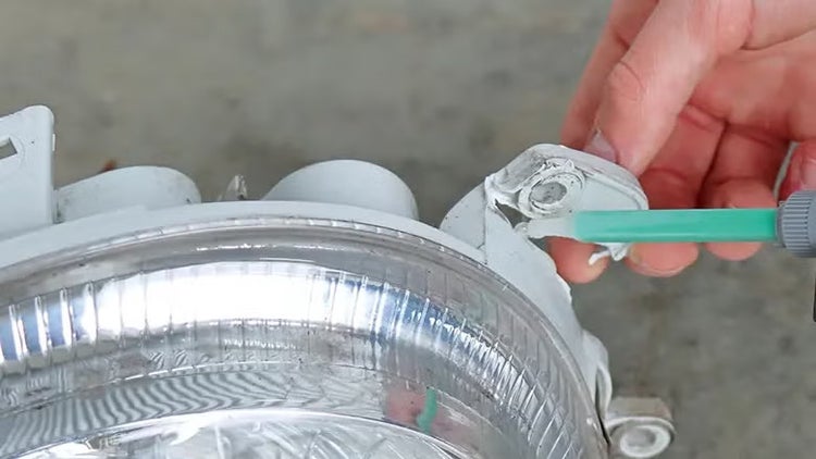 Eine Hand trägt Klebstoff mit einer Dosierspritze auf die Halterung eines Autoscheinwerfers auf, um eine Reparatur durchzuführen.