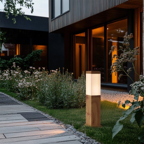 Moderne Wegeleuchte aus Holz in einem Garten neben einem gepflasterten Weg vor einem beleuchteten Wohnhaus bei Dämmerung.