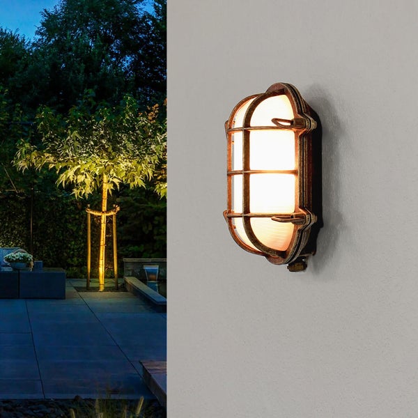 Außenansicht einer beleuchteten Schiffslampe an einer Wand, im Hintergrund ein Garten bei Nacht.