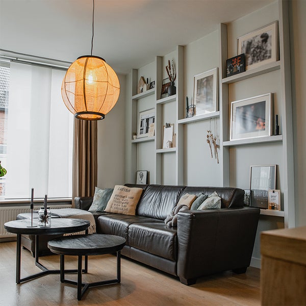Wohnzimmer mit dunklem Ledersofa, zwei runden Couchtischen, einer runden Pendelleuchte und einem eingebauten Wandregal mit Bilderrahmen.