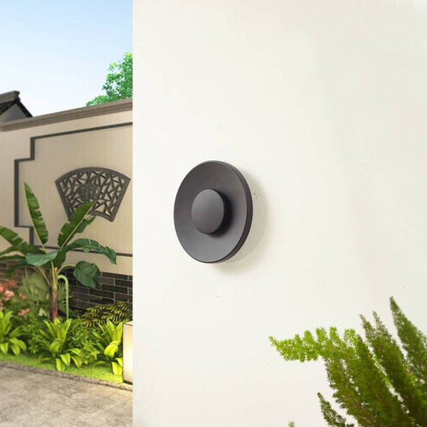 Moderne schwarze Außenwandleuchte in runder Form, montiert an einer hellen Fassade vor einem Garten mit grünen Pflanzen.