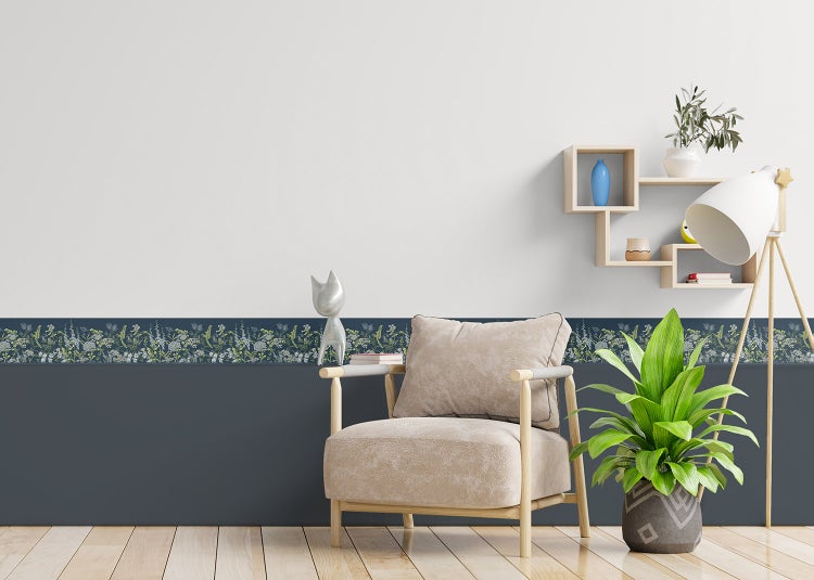Selbstklebende Tapetenbordüre mit Blumenmuster an einer Wand in einem hellen Zimmer mit Sessel, Holzregal und Zimmerpflanze.