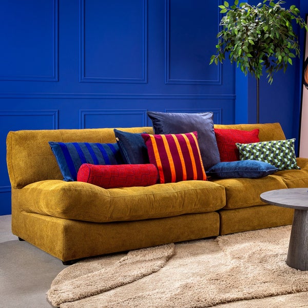 Modernes Wohnzimmer mit gelbem Polstersofa, verschiedenen Dekokissen, einem runden Teppich und einer Zimmerpflanze vor einer Wand mit Wandleisten.