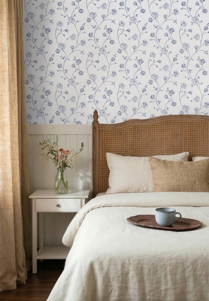 Helles Schlafzimmer mit blau geblümter Tapete, Bett mit Rattankopfteil, weißem Nachttisch, Glasvase und hellen Textilien.